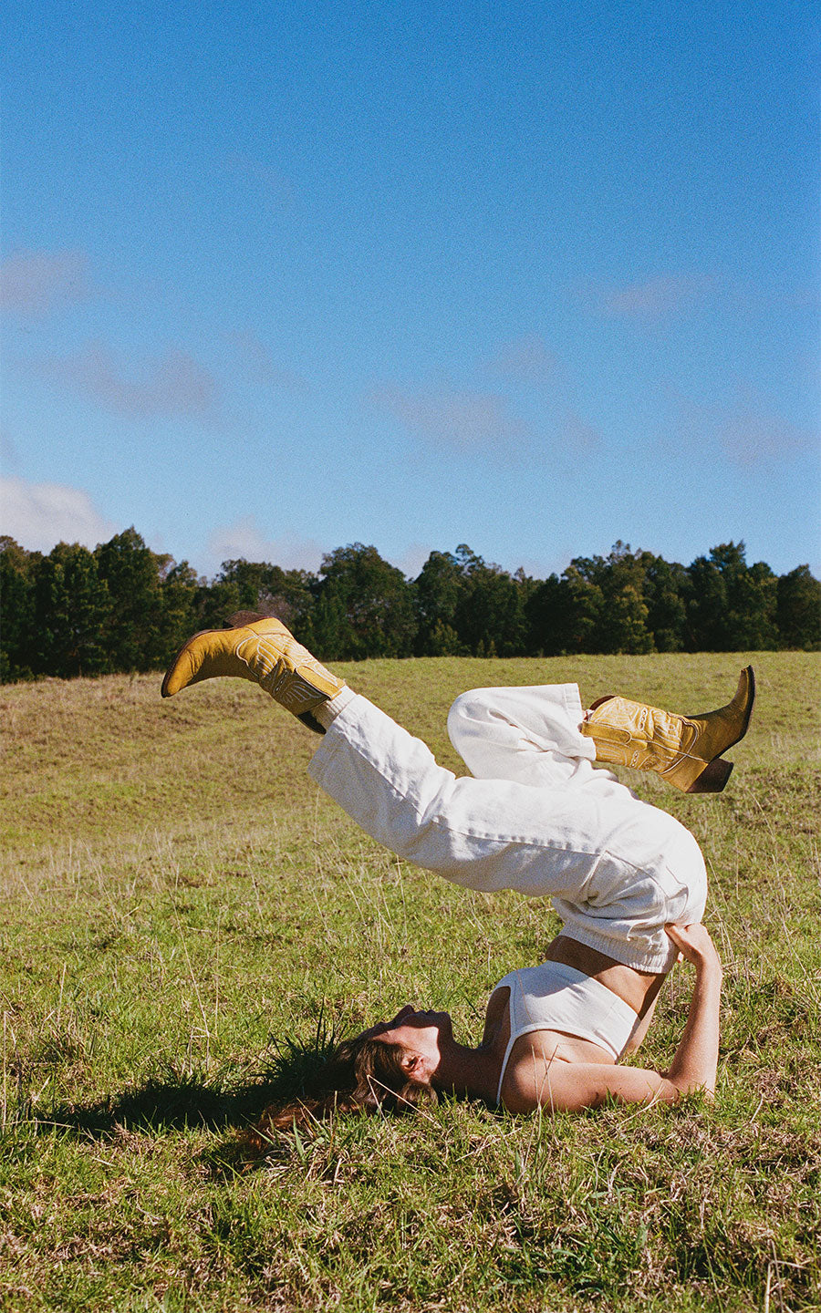 A woman lies on her back in a grassy field, wearing white pants, a white crop top, and gold cowboy boots.